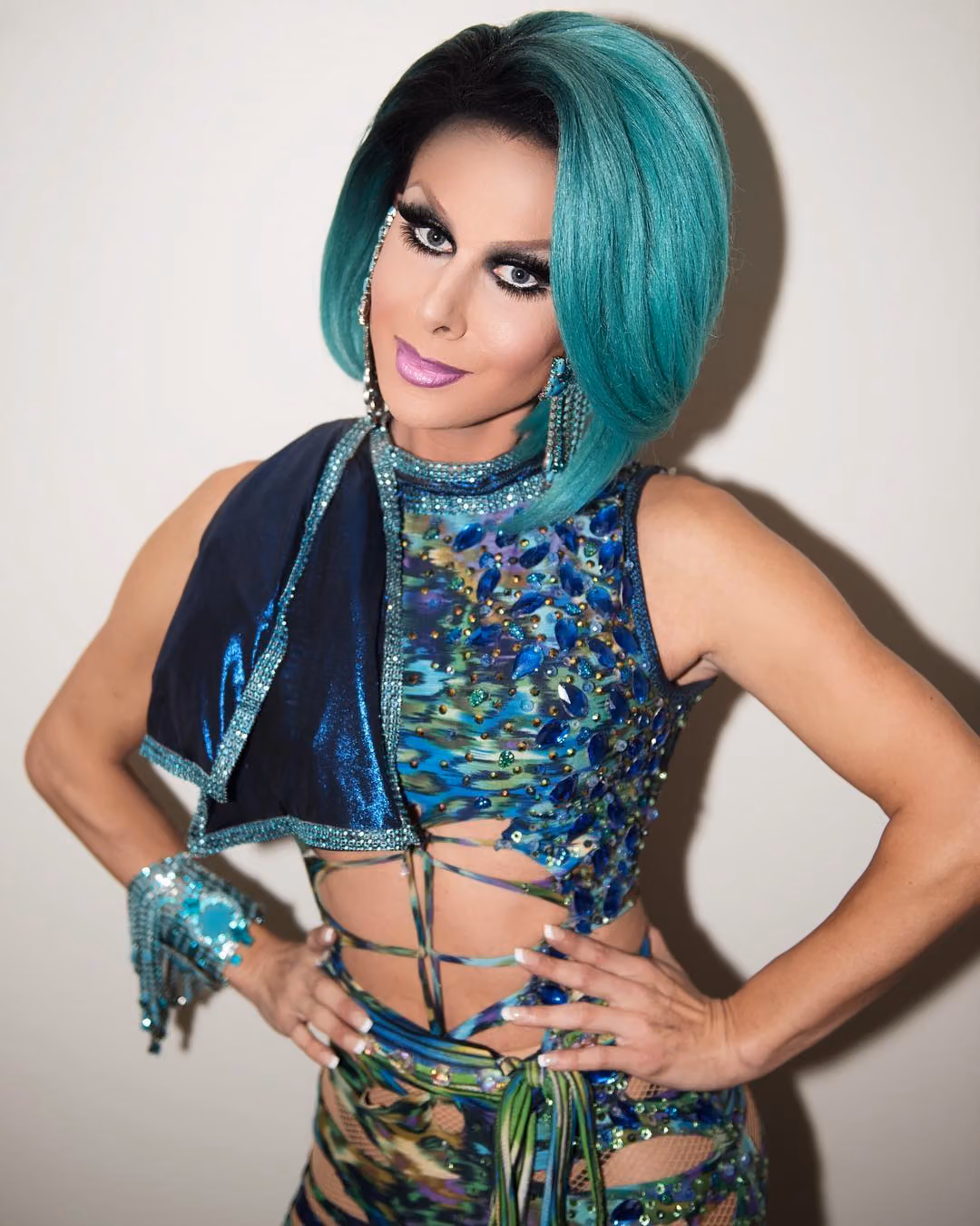 Drag queen with a teal bob wig in a jeweled peacock-print outfit posing with hands on hips