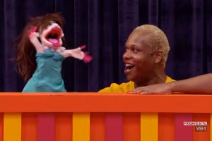 Contestant out of drag works a puppet in a teal dress over the striped puppet stage