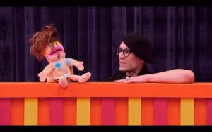 Contestant in glasses and a black beanie works a brown-haired puppet over the striped puppet stage