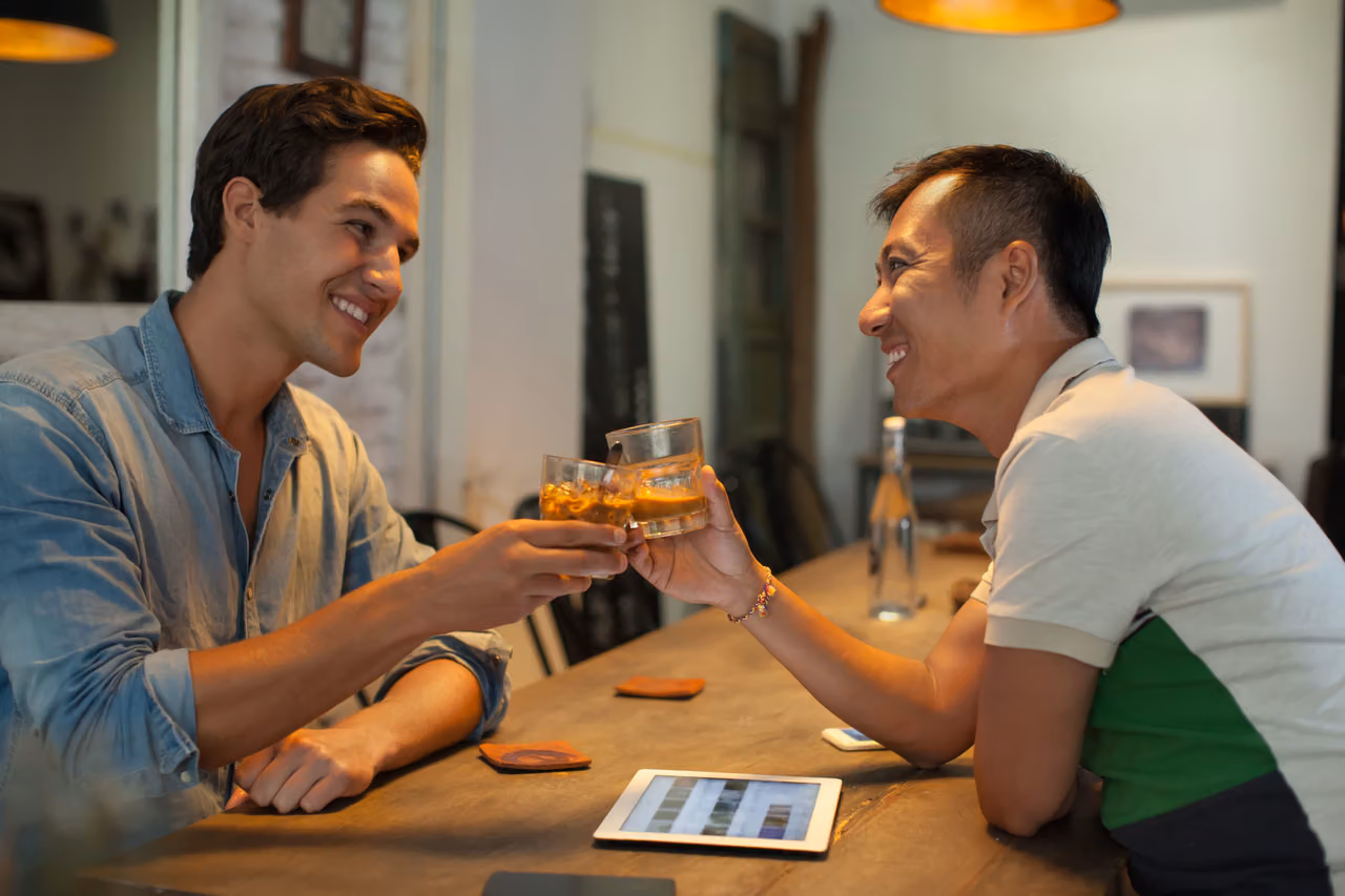 Two smiling men clinking iced drinks across a cafe table with a tablet between them