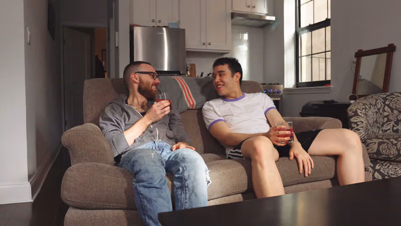 Still from The Art of Twinking: two men chat on a couch with drinks in an apartment living room