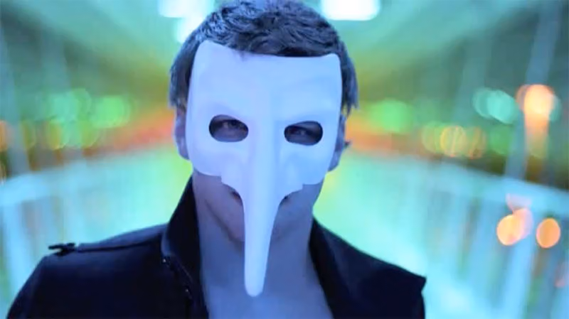 Man in a white long-nosed mask against blurred neon lights, one of the masked characters in Black Spark's films