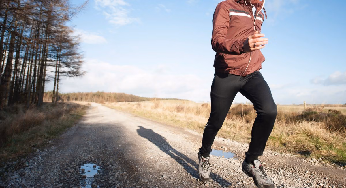 Runner in a maroon jacket and slim black training pants jogging down a gravel country road
