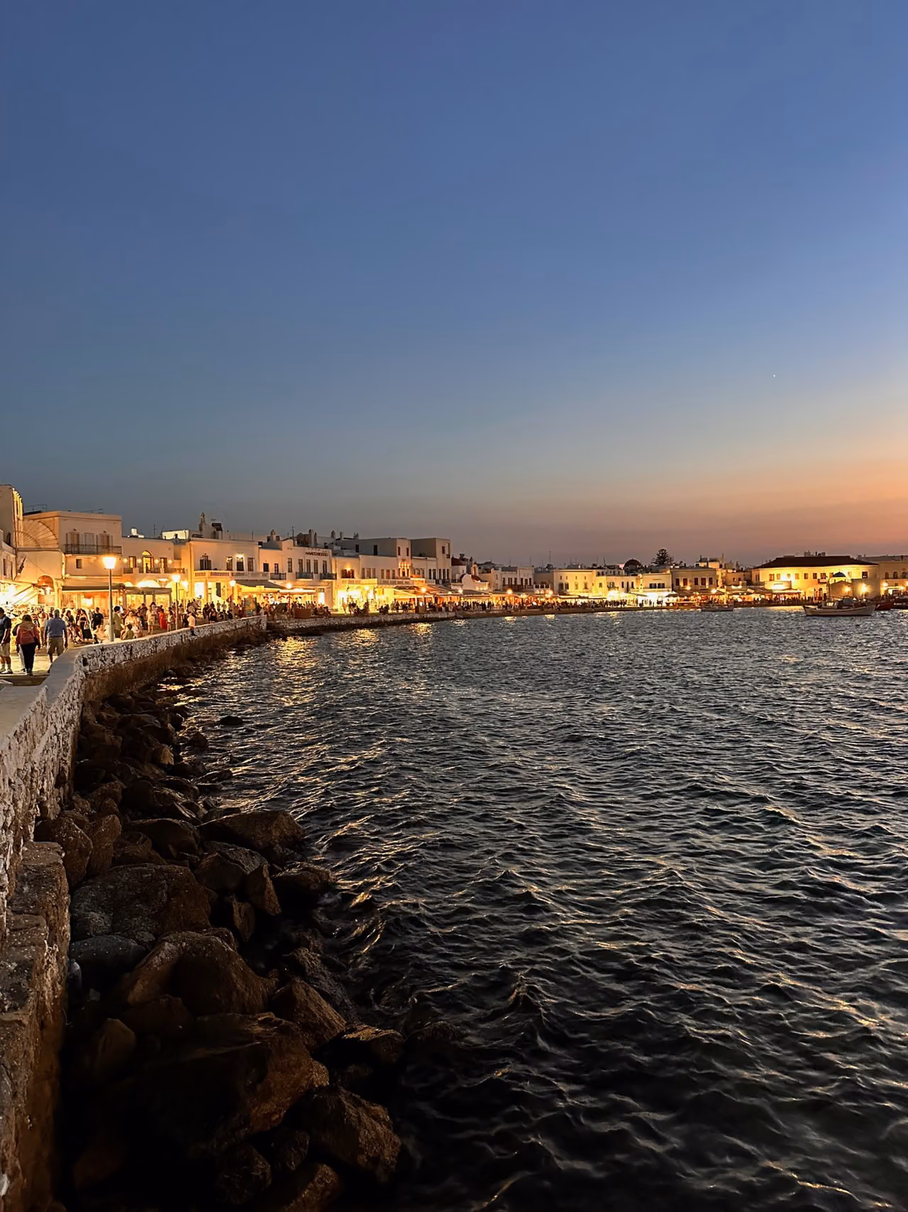 Illuminated restaurants and people along the Mykonos Town waterfront at dusk.