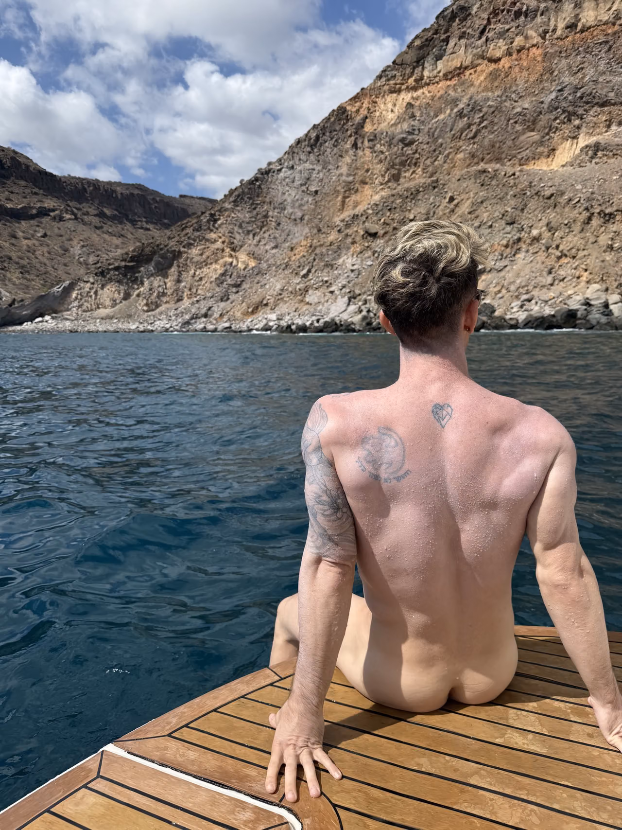 Mark sitting on the yacht’s swim platform facing a rocky cove, photographed from behind.