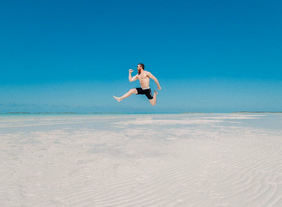 Shirtless man leaping midair over a white sandbar under a clear turquoise sky