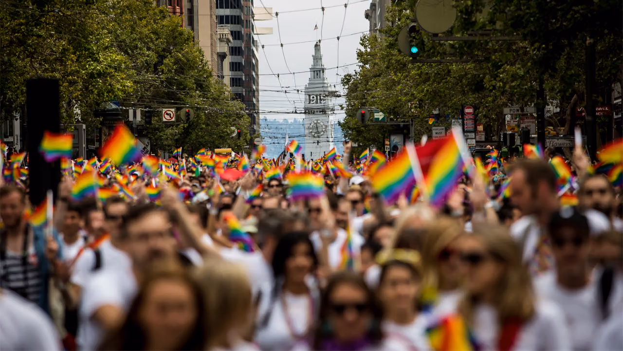 Crowd waving rainbow flags on Market Street at San Francisco Pride, with the Ferry Building ahead