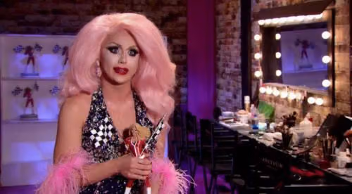 Farrah Moan in a big pink wig holding a doll in the Drag Race workroom