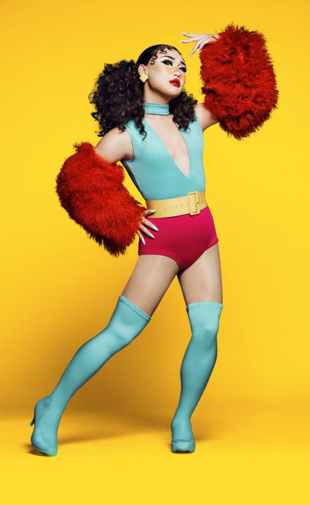 Soju in a teal plunging bodysuit, magenta briefs, red fur sleeves and teal thigh-high boots against a yellow backdrop