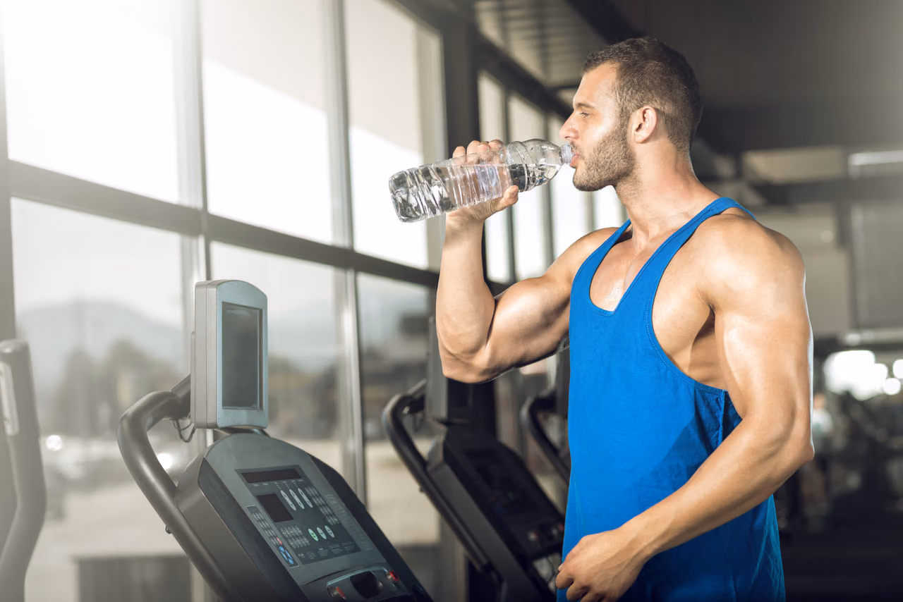 Muscular man in a blue tank top drinks from a water bottle beside a gym treadmill