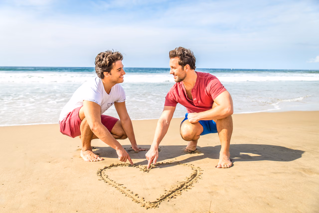 Two men smile at each other while drawing a heart in the sand on a beach