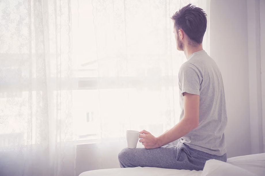 Man in a T-shirt and sweatpants sitting on a bed with a mug, looking out a bright window
