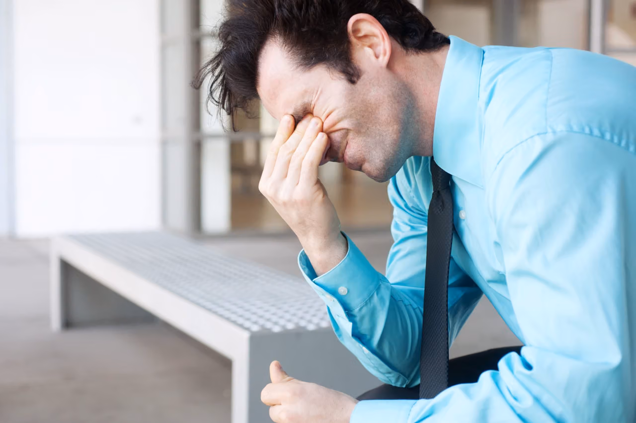 Man in a blue dress shirt and tie pinches the bridge of his nose in distress