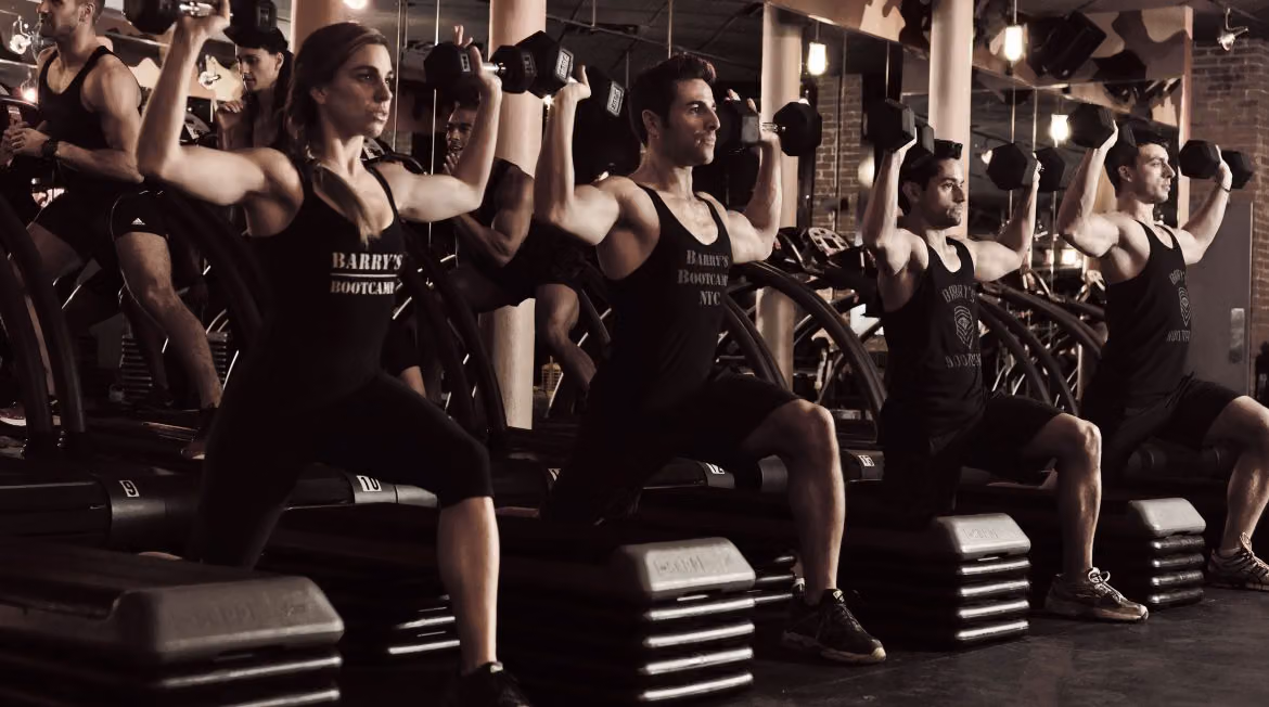 Barry's Bootcamp class in black tanks doing lunges with overhead dumbbell presses on step platforms