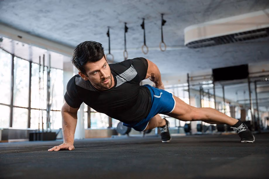Man in a black athletic tee and blue shorts doing a one-arm push-up in a gym