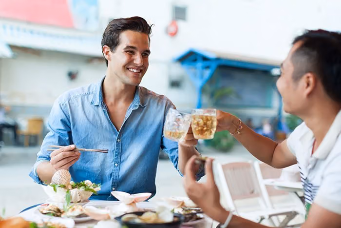 Two men toasting with glasses of beer over a seafood meal at an outdoor restaurant