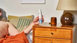 white man in bed reaching for lubricant bottle on wooden dresser