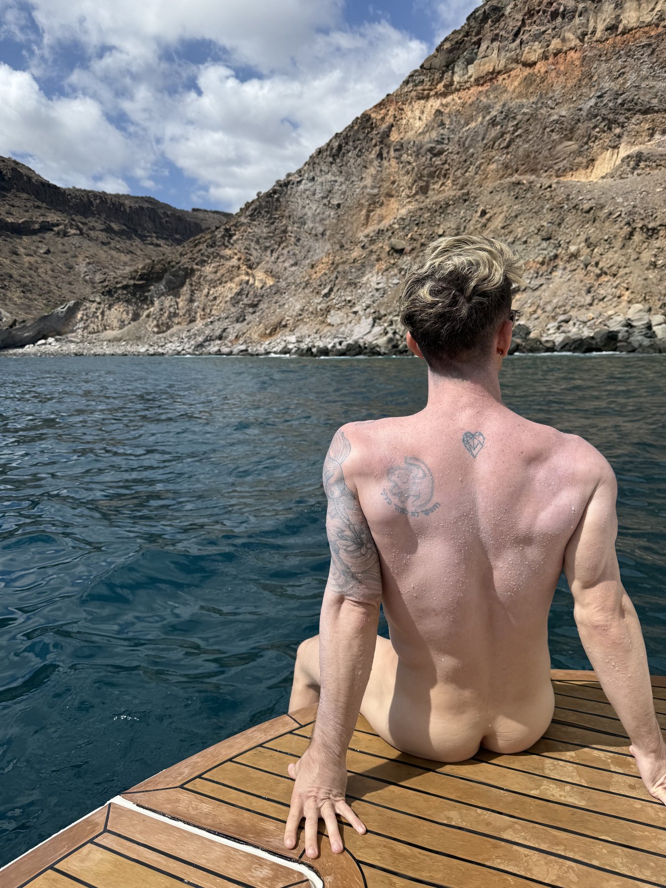 Mark sitting on the yacht’s swim platform facing a rocky cove, photographed from behind.