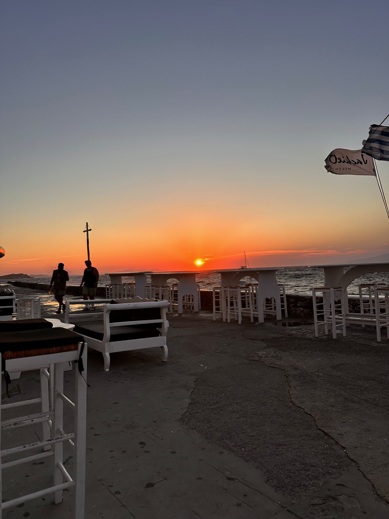 Orange sunset over the sea beside the Jackie O’s flag and waterfront seats.
