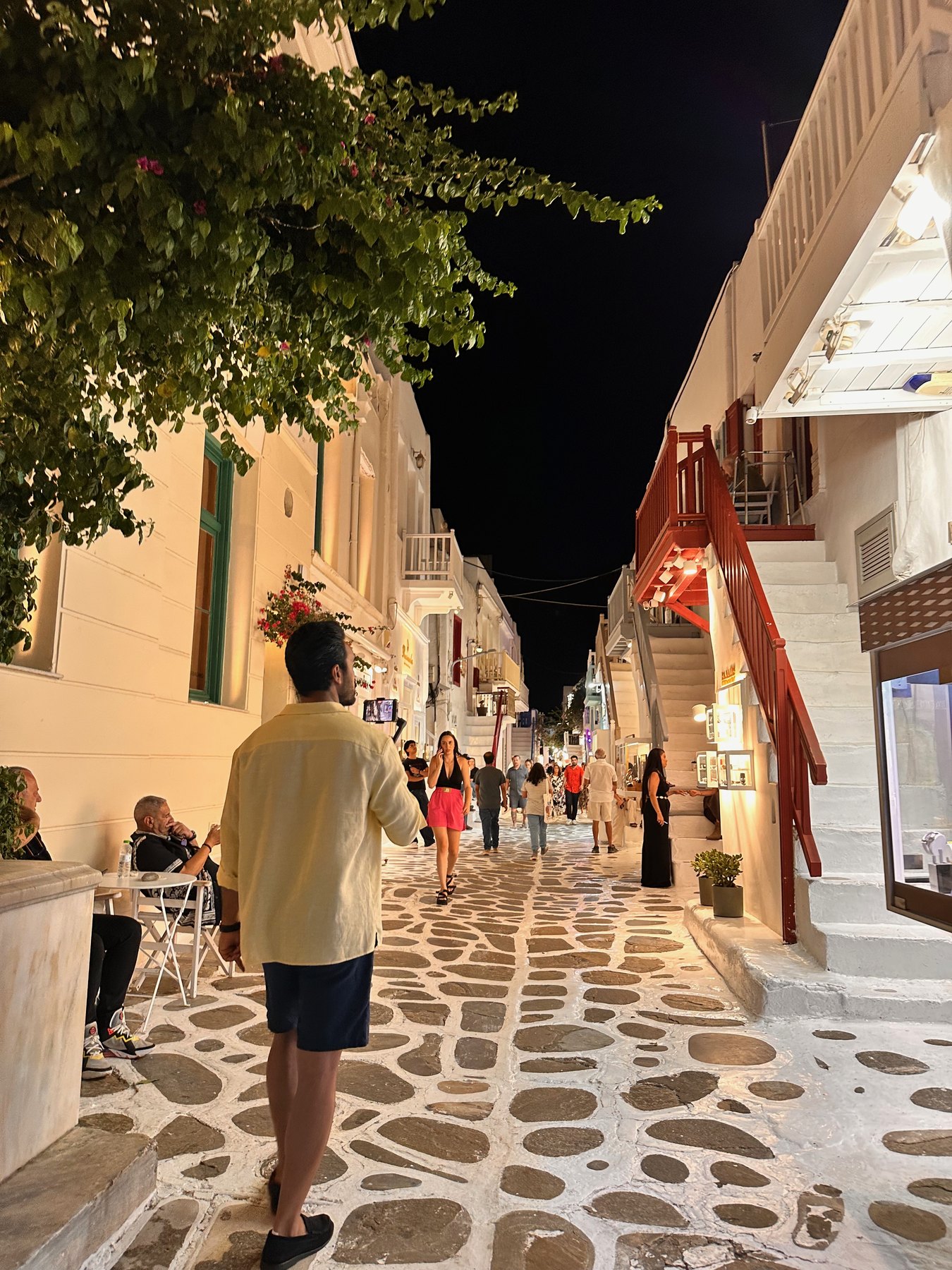 People walking between illuminated shops in a narrow Mykonos Town lane.