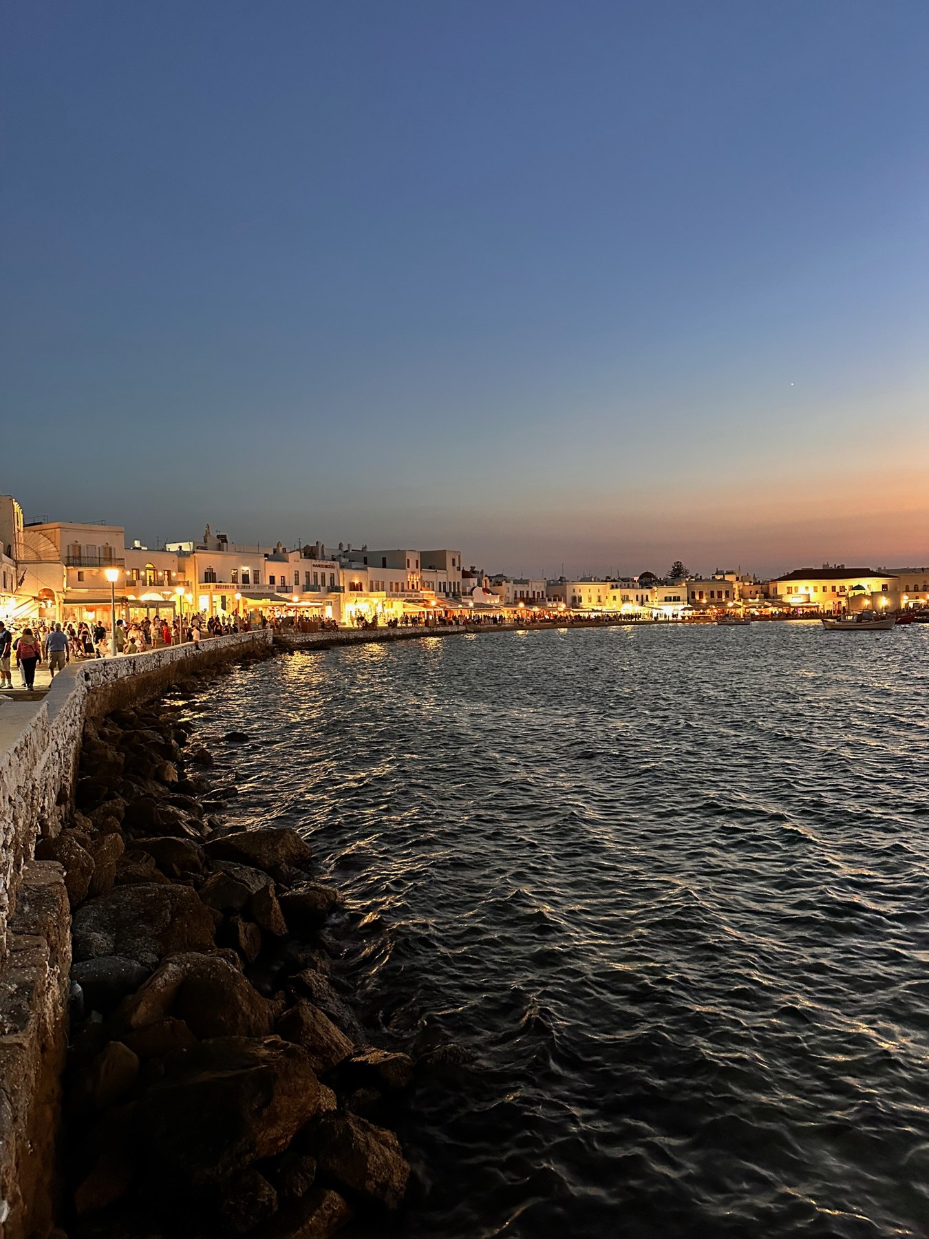 Illuminated restaurants and people along the Mykonos Town waterfront at dusk.